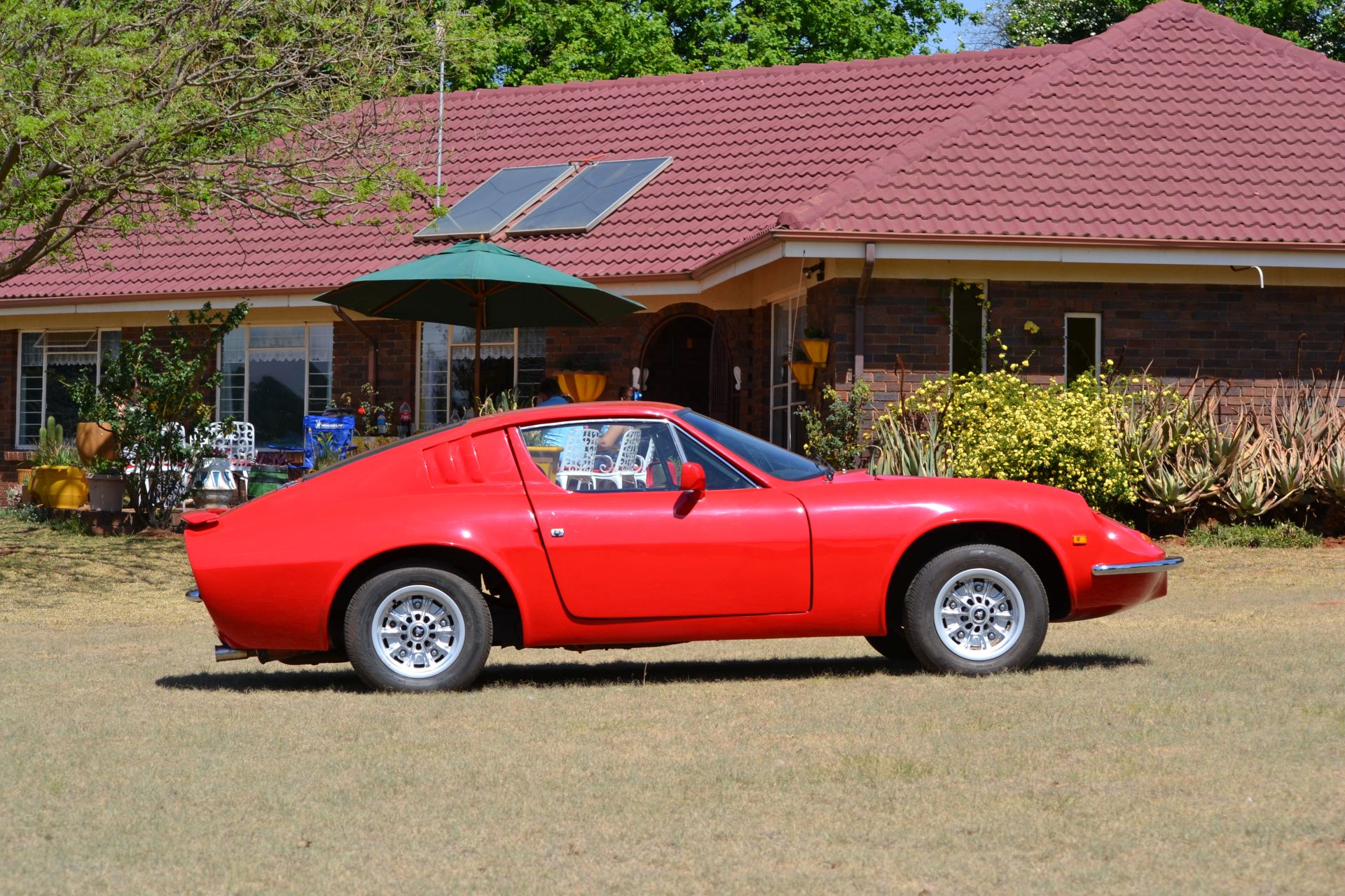 A red classic sports car parked on grass in front of a brick house. Click to visit vwpuma.co.uk.
